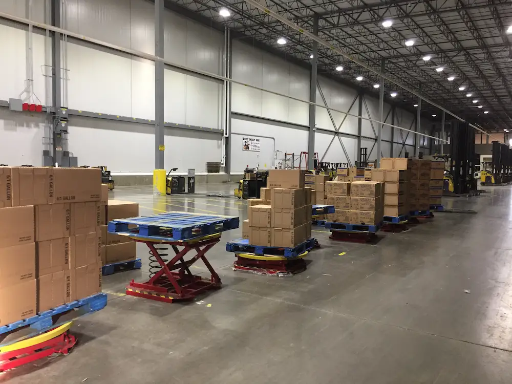 Warehouse with stacked cartons on blue pallets atop red pallet positioners; forklifts in background